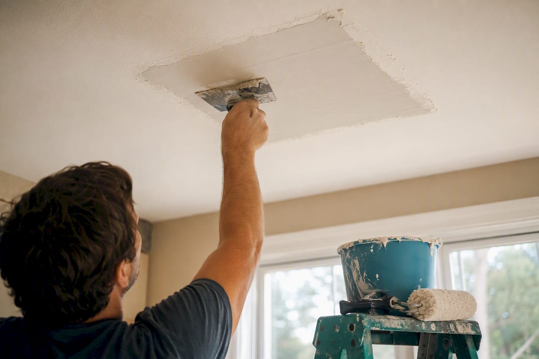 Hands applying joint compound to ceiling drywall patch